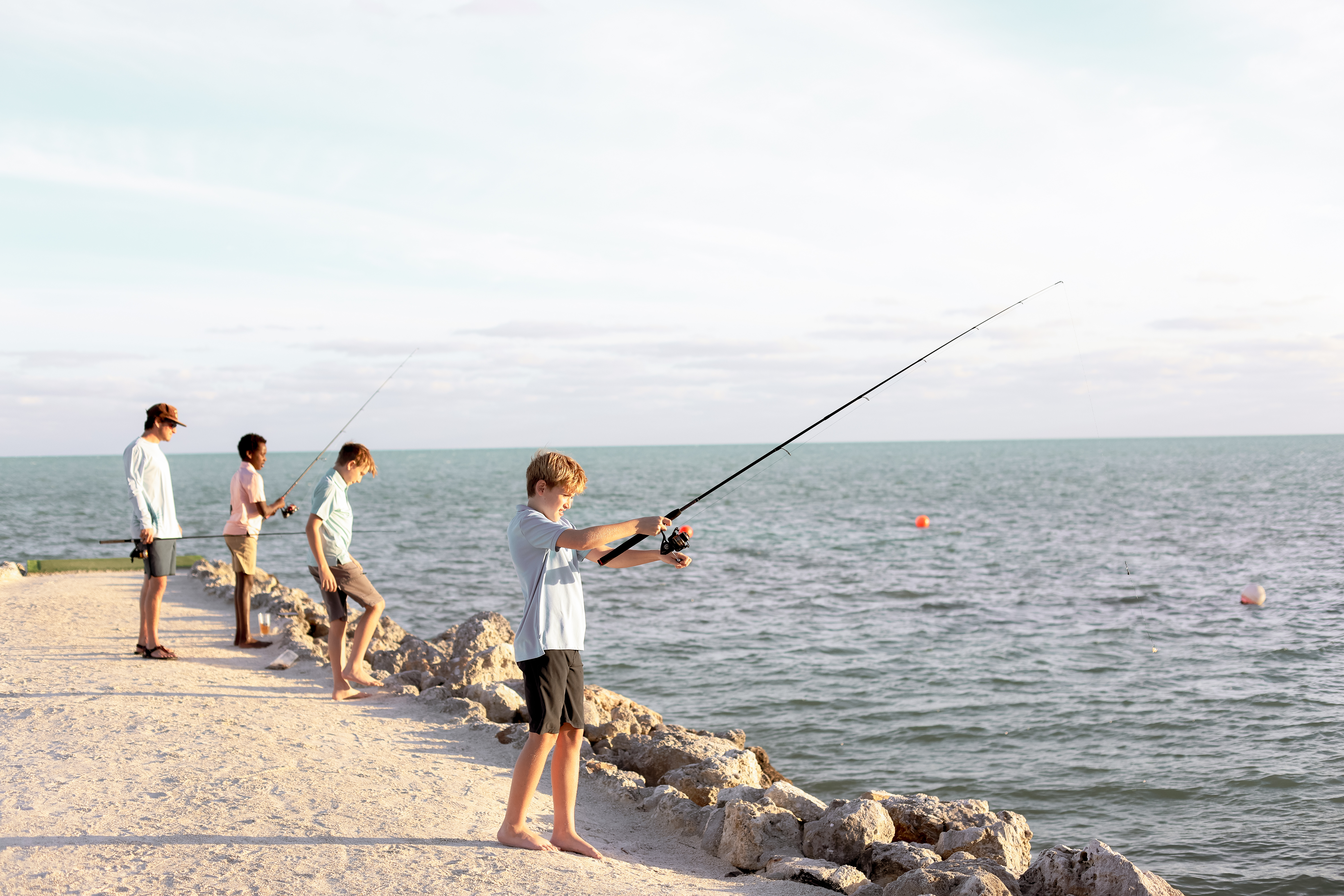Four people are fishing on a rocky shoreline by the sea, holding rods and looking towards the water.