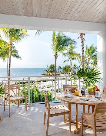 A patio with wooden furniture overlooks a pool and ocean, surrounded by palm trees on a sunny day.